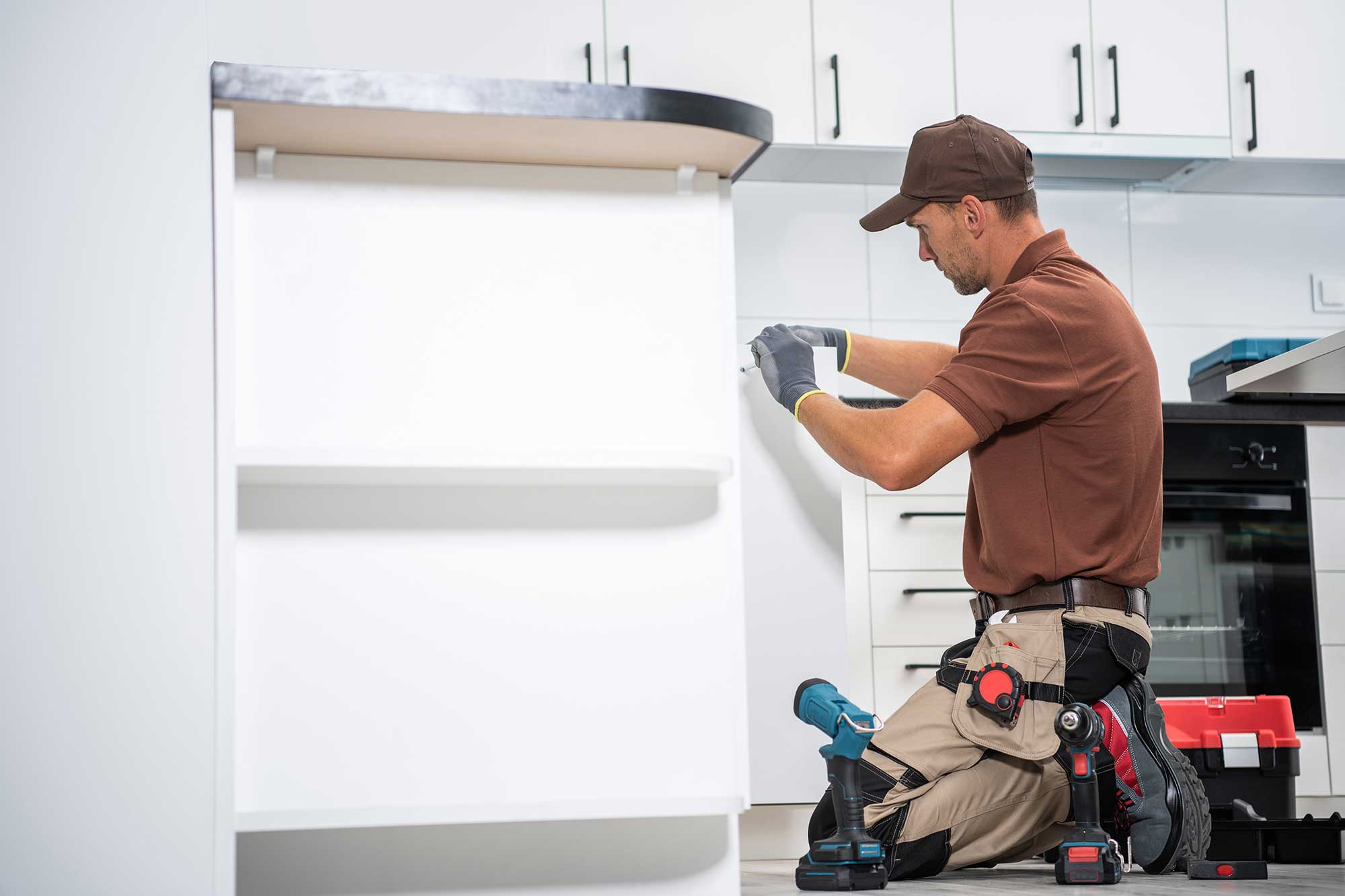 A man in a brown shirt and pants repairs a kitchen cabinet, focused on his task with tools in hand.