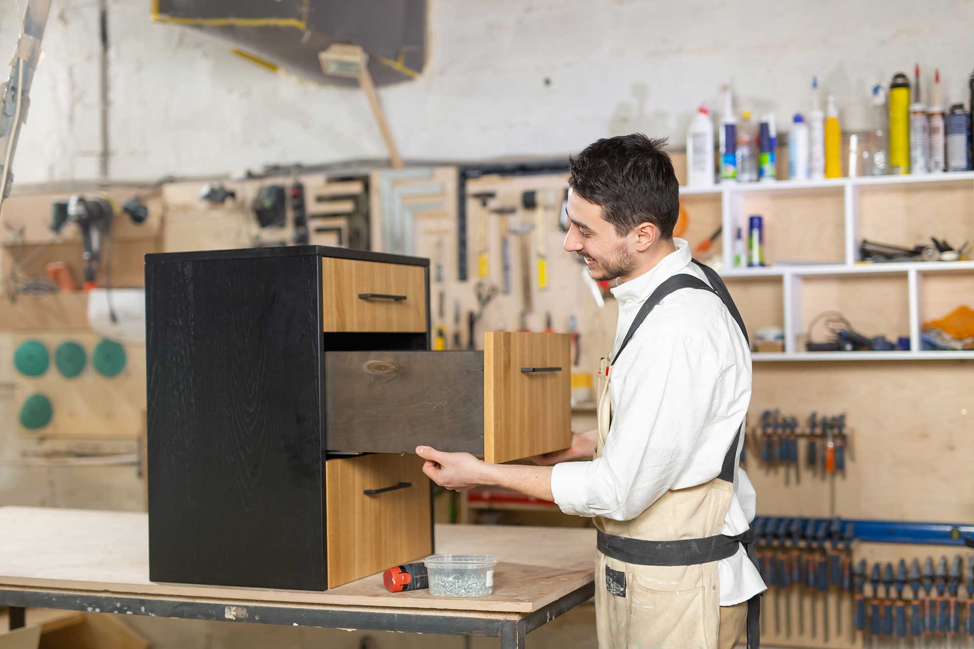 A man holds a wooden cabinet in a workshop, surrounded by tools and materials for woodworking.